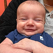 Matt is registered to the contest to win money with this photo: adult_partial, arms, baby, chubby_cheeks, close_up, cozy, hand, happy, holding, indoor, infant, jacket, necklace, onesie, orange_wall, person, portrait, skin, smile, wooden_chair