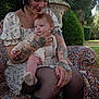 Lilith participe au concours pour gagner de l'argent avec cette photo : woman, baby, tattoos, outdoor, nature, greenery, floral_dress, child, holding, portrait, seated, patterned_chair, grass, trees, mother, infant, legs, skin, face, smile