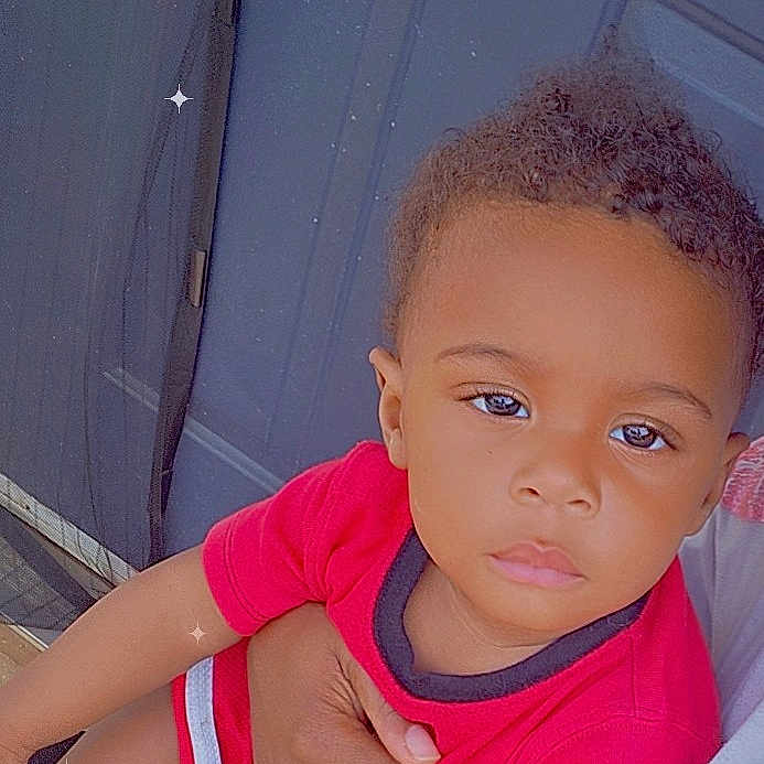 Ty'Zaiah is registered to the contest to win money with this photo: adult_hand, blue_door, casual, child, closeup, door, hair, hand, indoor, mesh_screen, person, portrait, red_clothing, serious_expression, shirt, shorts, sitting, skin, sparkles, toddler