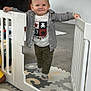 toddler, baby_gate, child, smiling, indoor, floor_mat, socks, shirt, pants, kitchen, modern, toy, play_area, person, holding, standing, white, floor, cute, happy