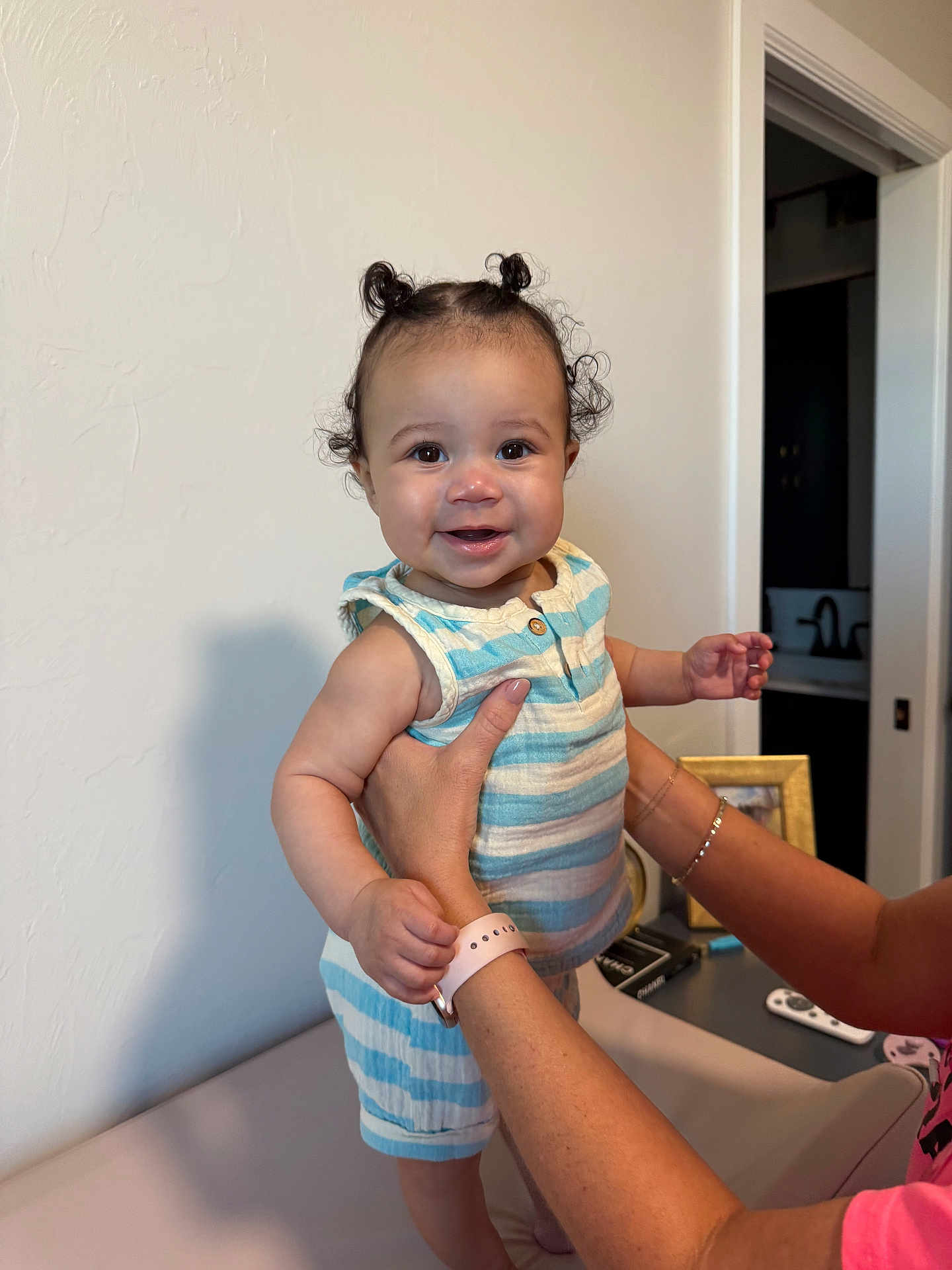 Jiovanni is registered to the contest to win money with this photo: baby, child, smile, curly_hair, striped_clothing, romper, hands, indoor, person, supporting_hands, face, cute, happy, standing, home, wall, picture_frame, remote_control, arm, skin
