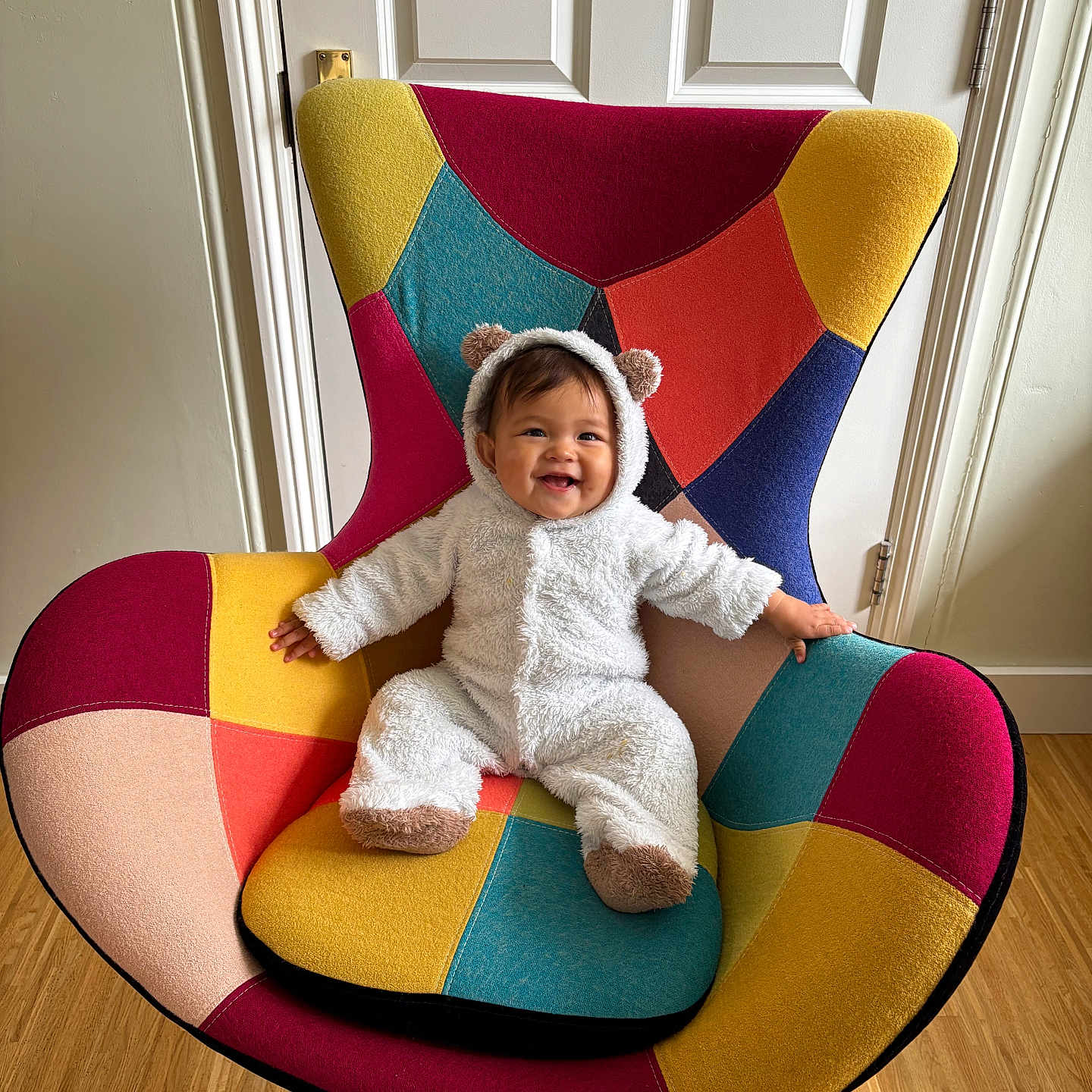 Isabella is registered to the contest to win money with this photo: baby, bear_costume, child, colorful_chair, cozy, cute, door, furniture, happy, home, indoor, onesie, patchwork, person, plush, portrait, seat, smiling, wooden_floor, young_child