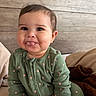 baby, child, tongue_out, green_onesie, hearts_pattern, sitting, bed, blanket, wooden_headboard, cute, infant, smiling, indoor, cozy, portrait, person, face, young_child, playful, comfortable