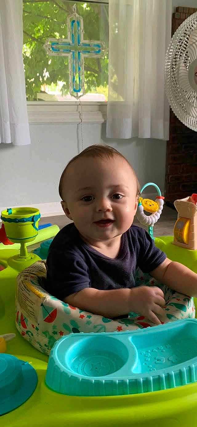 Jay is registered to the contest to win money with this photo: baby, child, indoor, activity_center, toys, smiling, face, curtains, window, cross, fan, furniture, person, colorful, seat, play, happy, young_child, infant, home