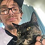 Ivy joined the competition — help win amazing prizes! person, cat, glasses, cap, tortoiseshell_cat, yellow_eyes, hand, ring, earrings, curtains, indoor, closeup, portrait, facial_hair, black_clothing, natural_light, whiskers, feline, pet, holding