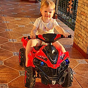 Daria joined the competition — help win amazing prizes! baby, child, cute, daylight, hands, outdoor, person, play, porch, red, ride, short_hair, small, smile, sunlight, tile_floor, toddler, toy_atv, vehicle, white_shirt