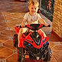 baby, child, cute, daylight, hands, outdoor, person, play, porch, red, ride, short_hair, small, smile, sunlight, tile_floor, toddler, toy_atv, vehicle, white_shirt