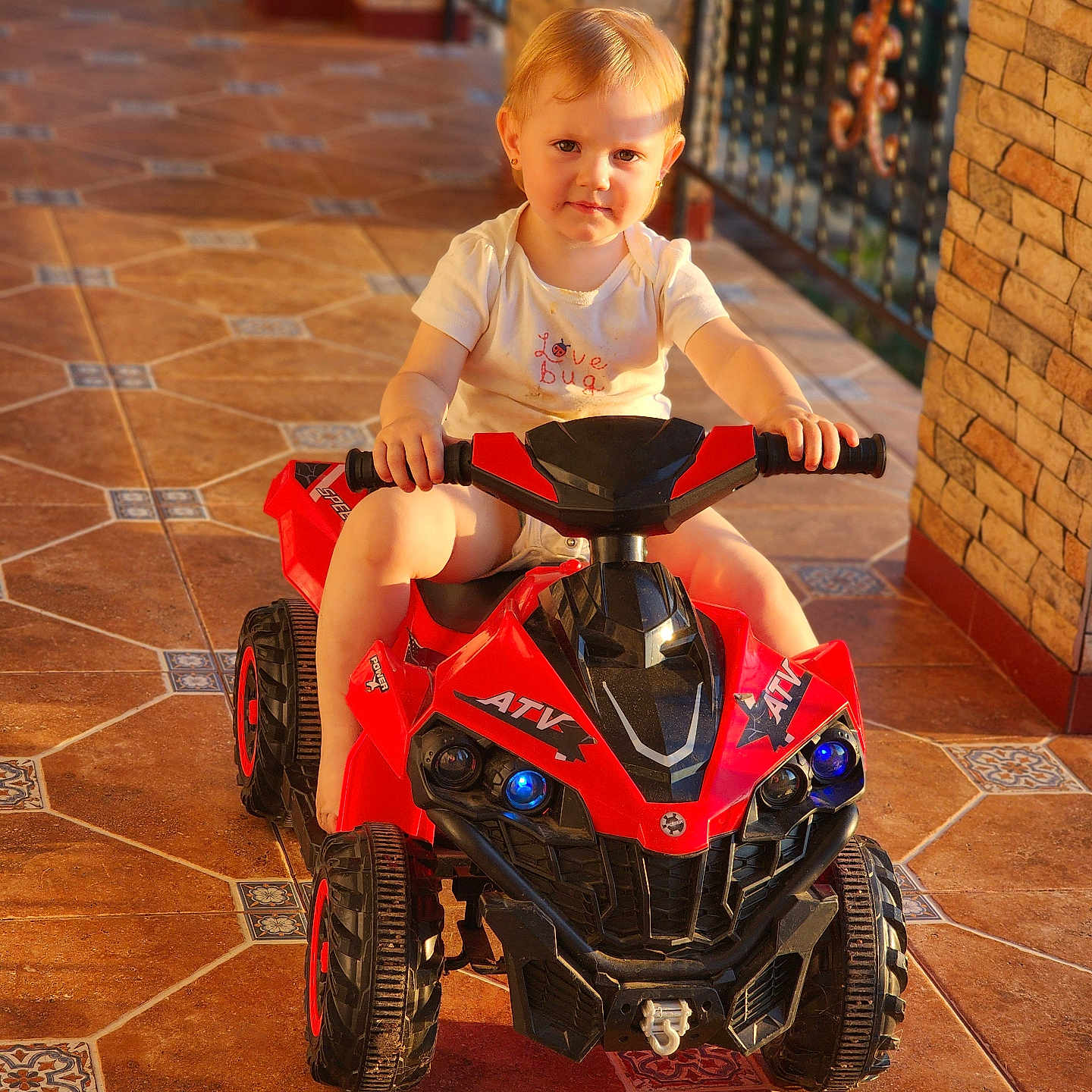 Daria joined the competition — help win amazing prizes! baby, child, cute, daylight, hands, outdoor, person, play, porch, red, ride, short_hair, small, smile, sunlight, tile_floor, toddler, toy_atv, vehicle, white_shirt