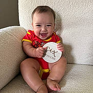 Kai joined the competition — help win amazing prizes! baby, child, infant, red_onesie, smiling, happy, couch, furniture, sign, wooden_sign, text, six_months, sitting, indoors, cute, portrait, skin, person, face, legs