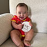 baby, child, infant, red_onesie, smiling, happy, couch, furniture, sign, wooden_sign, text, six_months, sitting, indoors, cute, portrait, skin, person, face, legs