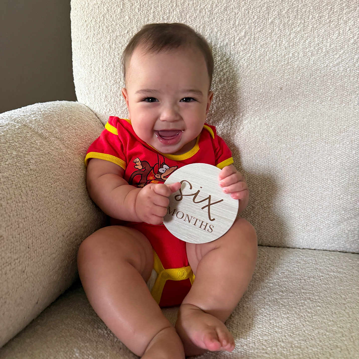 Kai joined the competition — help win amazing prizes! baby, child, infant, red_onesie, smiling, happy, couch, furniture, sign, wooden_sign, text, six_months, sitting, indoors, cute, portrait, skin, person, face, legs