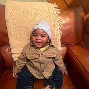 Levi is registered to the contest to win money with this photo: baby, child, toddler, smile, hat, jacket, denim_pants, leather_chair, blanket, happy, indoors, person, seated, cute, portrait, cozy, young_child, fashion, casual, warm_lighting