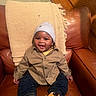baby, child, toddler, smile, hat, jacket, denim_pants, leather_chair, blanket, happy, indoors, person, seated, cute, portrait, cozy, young_child, fashion, casual, warm_lighting