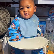 Levi is registered to the contest to win money with this photo: baby, child, high_chair, bib, pajamas, indoor, kitchen, fan, utensils, plate, face, person, cute, infant, tableware, cabinet, countertop, expression, sitting, clothing