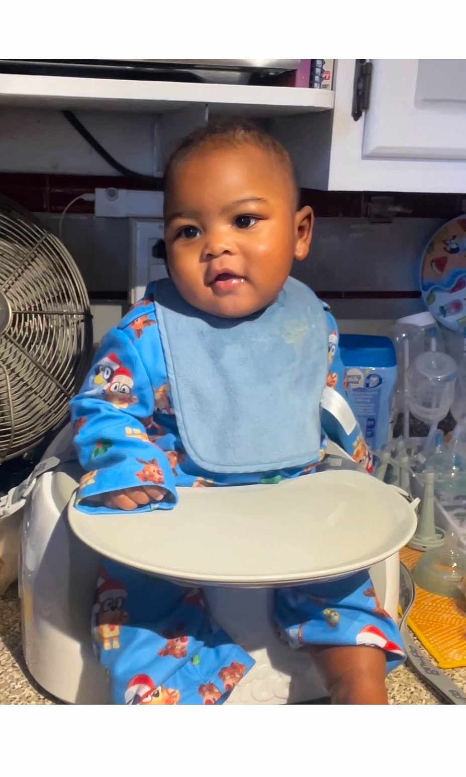Levi is registered to the contest to win money with this photo: baby, child, high_chair, bib, pajamas, indoor, kitchen, fan, utensils, plate, face, person, cute, infant, tableware, cabinet, countertop, expression, sitting, clothing