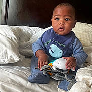 Levi is registered to the contest to win money with this photo: baby, child, infant, toy, bed, pillow, blue_clothing, bib, shark_design, hand, face, eyes, fingers, footwear, indoor, soft_lighting, wooden_headboard, curious_expression, sitting, crab_toy