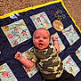 baby, infant, child, quilt, carpet, camouflage_shirt, black_pants, lying_down, smiling, happy, cute, indoor, colorful, cartoon_characters, fabric, pattern, person, portrait, young, playful