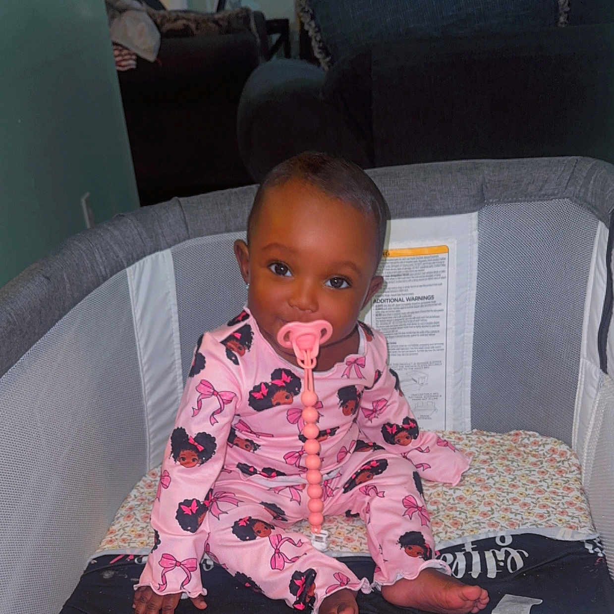 Melody is registered to the contest to win money with this photo: baby, child, pacifier, pink_pajamas, playpen, infant, sitting, cute, indoors, blanket, face, foot, hand, toy, clothing, furniture, living_room, expression, person, portrait