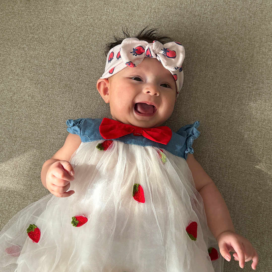 Luna joined the competition — help win amazing prizes! baby, child, smiling, dress, headband, strawberry, red_bow, happy, indoor, couch, fabric, cute, infant, person, apparel, childhood, lying_down, soft_texture, portrait, clothing