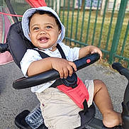 Kais a rejoint le concours — aidez-le/la à gagner de superbes lots ! toddler, child, smiling, happy, outdoor, tricycle, hat, shorts, white_shirt, sitting, handlebar, playground, sunny_day, fence, water_bottle, footwear, baby, seatbelt, toy, recreation