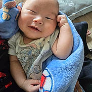 Jaylen joined the competition — help win amazing prizes! baby, infant, child, portrait, sleepy, smiling, face, eyes, hand, blanket, blue_blanket, cartoon_clothing, onesie, sitting, indoors, couch, pillow, cozy, close_up, relaxed