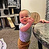 baby, child, standing, indoor, carpet, cat, cat_tree, milestone, plaque, table, clothing, pants, shirt, hand, face, head, smiling, floor, furniture, curtain