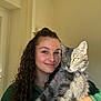 woman, cat, pet, curly_hair, green_sweater, indoor, portrait, smile, holding, gray_tabby, sunlight, yellow_eyes, face, feline, happy, human, long_hair, animal, cozy, casual