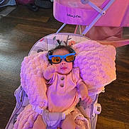 Layla is registered to the contest to win money with this photo: baby, sunglasses, blanket, baby_seat, indoor, wooden_floor, pink, cushion, infant, clothing, person, toddler, headwear, smiling, cute, child, playful, seatbelt, hair, toy