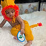 baby, infant, child, knitted_clothing, lion_hat, yellow, orange, plush_rug, card, months, indoor, playpen, animal_prints, cute, sitting, portrait, young_child, soft_texture, hand, feet