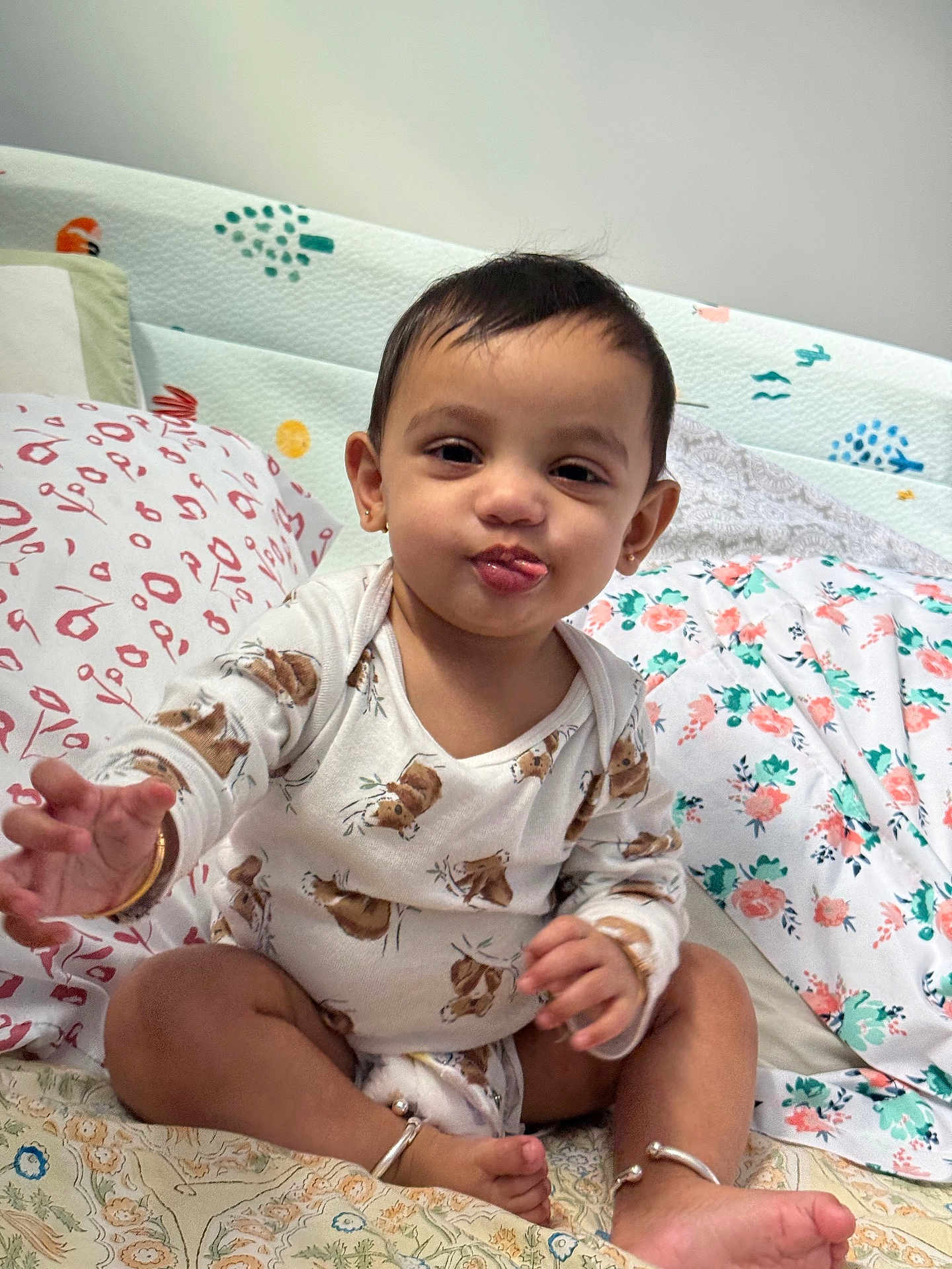 Vishruth is registered to the contest to win money with this photo: baby, child, infant, onesie, bed, pillow, floral_pattern, animal_print, bracelet, anklet, cute, tongue_out, smiling, indoor, person, sitting, hand, foot, face, playful