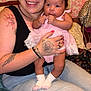 woman, baby, infant, tattoo, smile, holding, pink_dress, socks, indoor, bed, pillow, person, toddler, happy, casual, hair, face, jeans, hand, candid