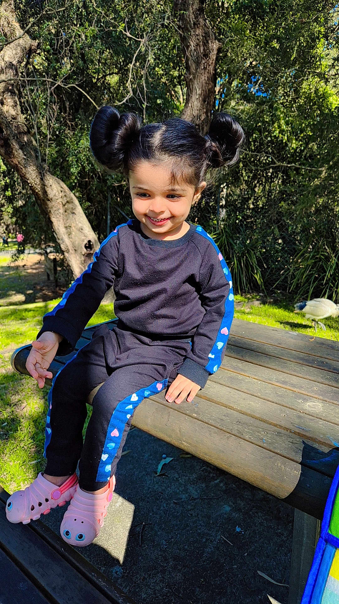 Nyrah is registered to the contest to win money with this photo: child, smiling, outdoor, park, bench, trees, sunlight, black_clothing, blue_stripes, pink_shoes, grass, bird, hair_buns, happy, person, casual_wear, nature, daylight, cute, playful