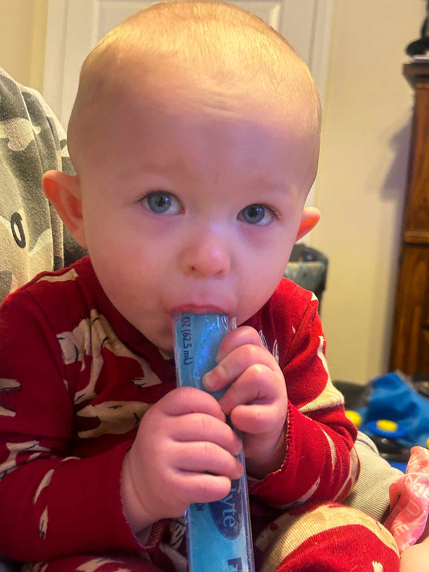 Kayden joined the competition — help win amazing prizes! toddler, child, pajamas, indoors, freeze_pop, hand, face, blue, red, toy, furniture, curious, closeup, person, chewing, baby, comfort, warm_clothing, cute, expression