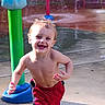toddler, child, smiling, running, water_park, splash_pad, wet, hair, red_shorts, bare_chest, outdoor, daylight, playground, fun, joy, water_spray, green_pole, blue_base, concrete, summer