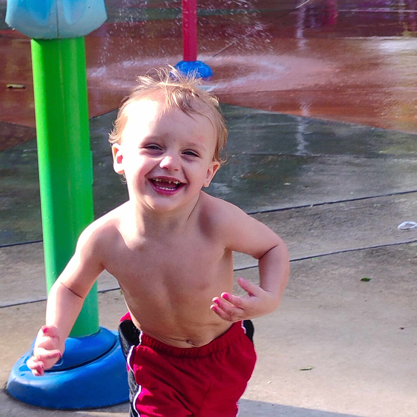 Alexander Hughes is registered to the contest to win money with this photo: toddler, child, smiling, running, water_park, splash_pad, wet, hair, red_shorts, bare_chest, outdoor, daylight, playground, fun, joy, water_spray, green_pole, blue_base, concrete, summer