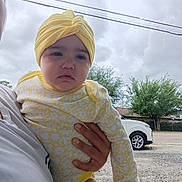 Camilla is registered to the contest to win money with this photo: baby, car, casual, child, clothing, cloudy_sky, daylight, expressionless, gravel, hand, headwear, holding, infant, nature, outdoor, person, road, tree, yellow_onesie, yellow_turban