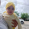 baby, car, casual, child, clothing, cloudy_sky, daylight, expressionless, gravel, hand, headwear, holding, infant, nature, outdoor, person, road, tree, yellow_onesie, yellow_turban