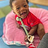 baby, child, smiling, happy, portrait, red_outfit, pink_seat, toy, teething_toy, indoor, sitting, infant, dark_skin, curly_hair, face, eyes, open_mouth, colorful_background, close_up, family_photo