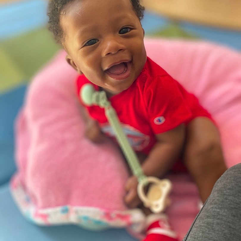 Axxel joined the competition — help win amazing prizes! baby, child, smiling, happy, portrait, red_outfit, pink_seat, toy, teething_toy, indoor, sitting, infant, dark_skin, curly_hair, face, eyes, open_mouth, colorful_background, close_up, family_photo