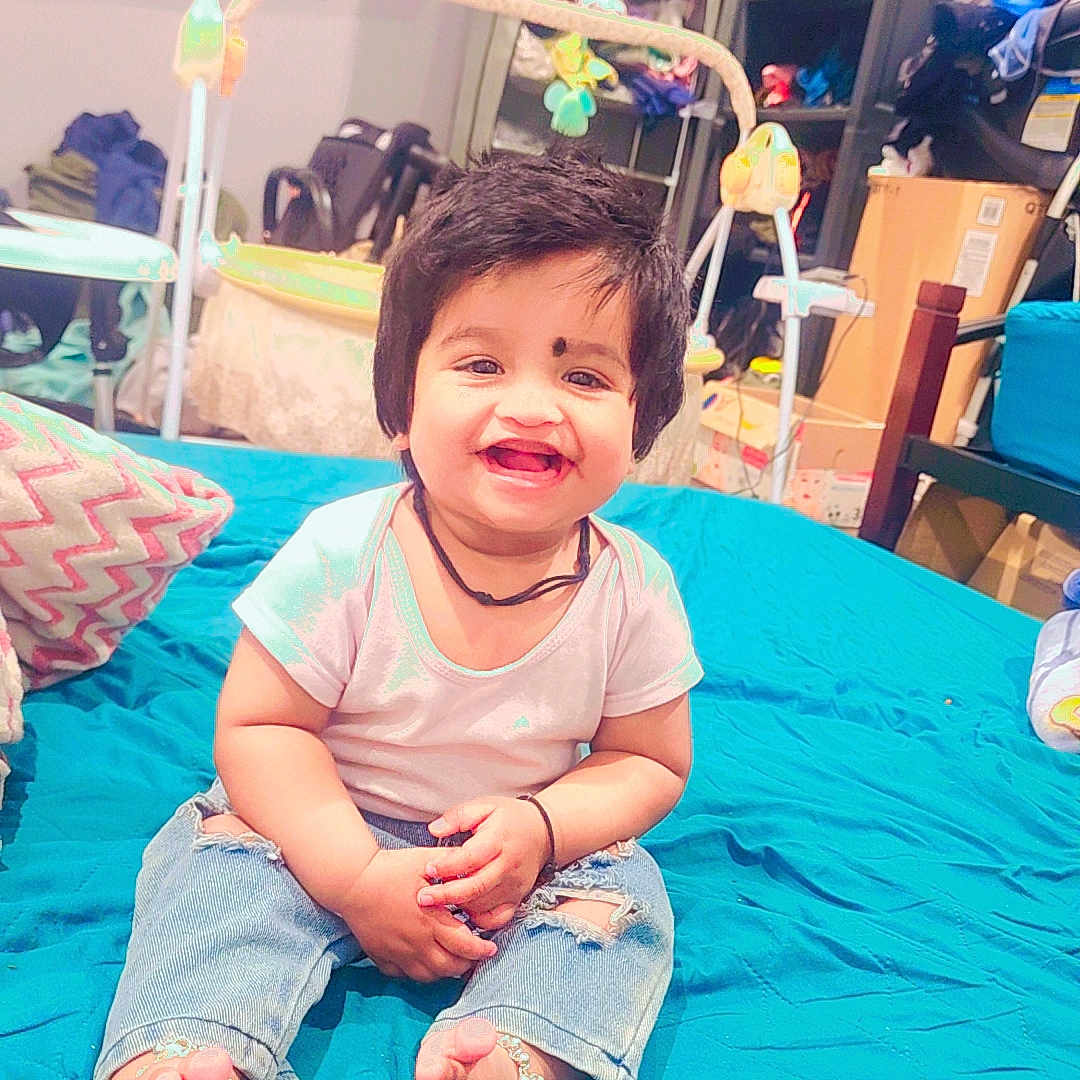 Keny is registered to the contest to win money with this photo: anklets, bed, blue_bedspread, casual_wear, child, clothes, footwear, furniture, hair, happy, indoor, jeans, person, room, shelf, smiling, television, toddler, toys, white_tshirt