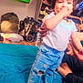 adult, barefoot, bed, blue_bedspread, child, clothing, furniture, hair_tie, hand, home, indoor, jeans, person, shelf, standing, television, thumb_sucking, toddler, toy, white_shirt