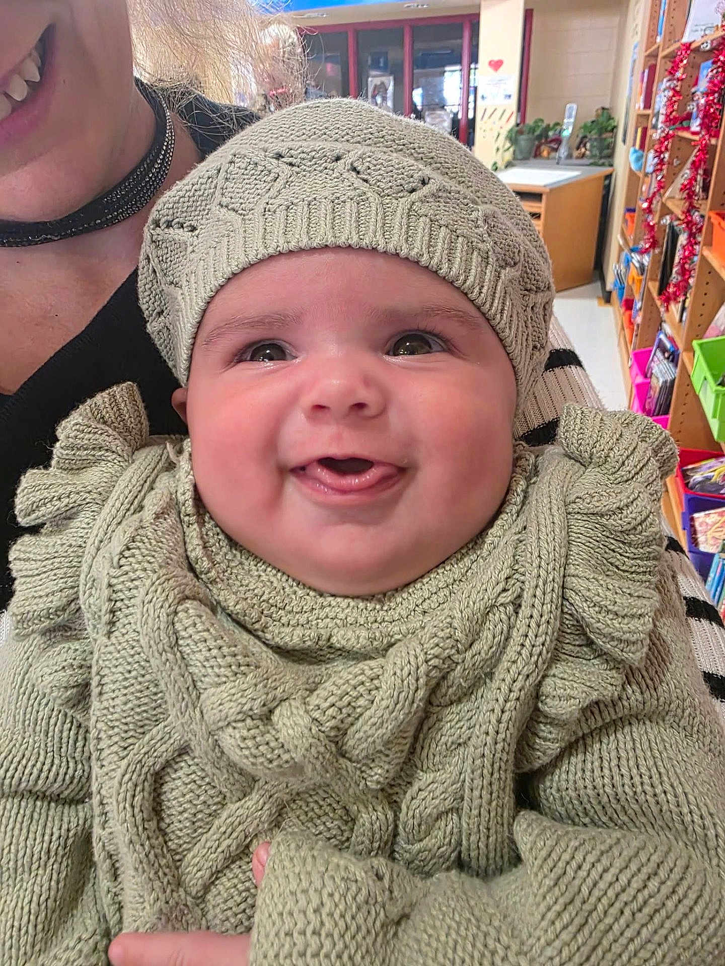 Lilah joined the competition — help win amazing prizes! baby, smiling, knitted_clothing, hat, indoor, person, adult, child, happy, closeup, face, cheeks, warm_clothing, sweater, holding, furniture, shelf, decorations, colorful, portrait