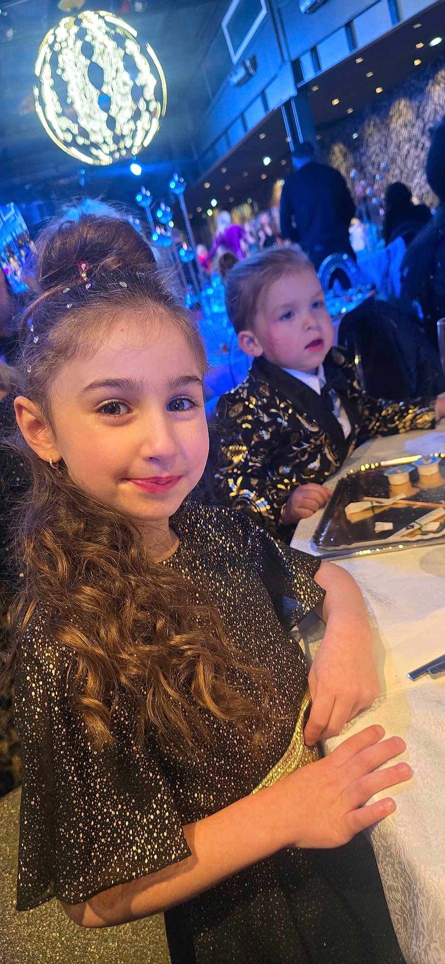 Lyna participe au concours pour gagner de l'argent avec cette photo : child, girl, boy, party, event, dress, glitter, curly_hair, table, food_tray, indoor, people, celebration, formal_wear, smile, hairstyle, lighting, decor, seated, background