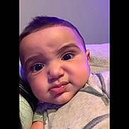 Ali is registered to the contest to win money with this photo: baby, infant, child, portrait, face, eyes, serious_expression, close_up, sitting, indoor, cushion, sofa, clothing, striped_shirt, purple_lighting, cozy, head, cheeks, photography, home