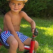 Isaac a rejoint le concours — aidez-le/la à gagner de superbes lots ! child, boy, smile, hat, straw_hat, tricycle, outdoor, grass, greenery, summer, shorts, striped_shorts, sunlight, play, toy, happy, portrait, bare_chest, nature, casual