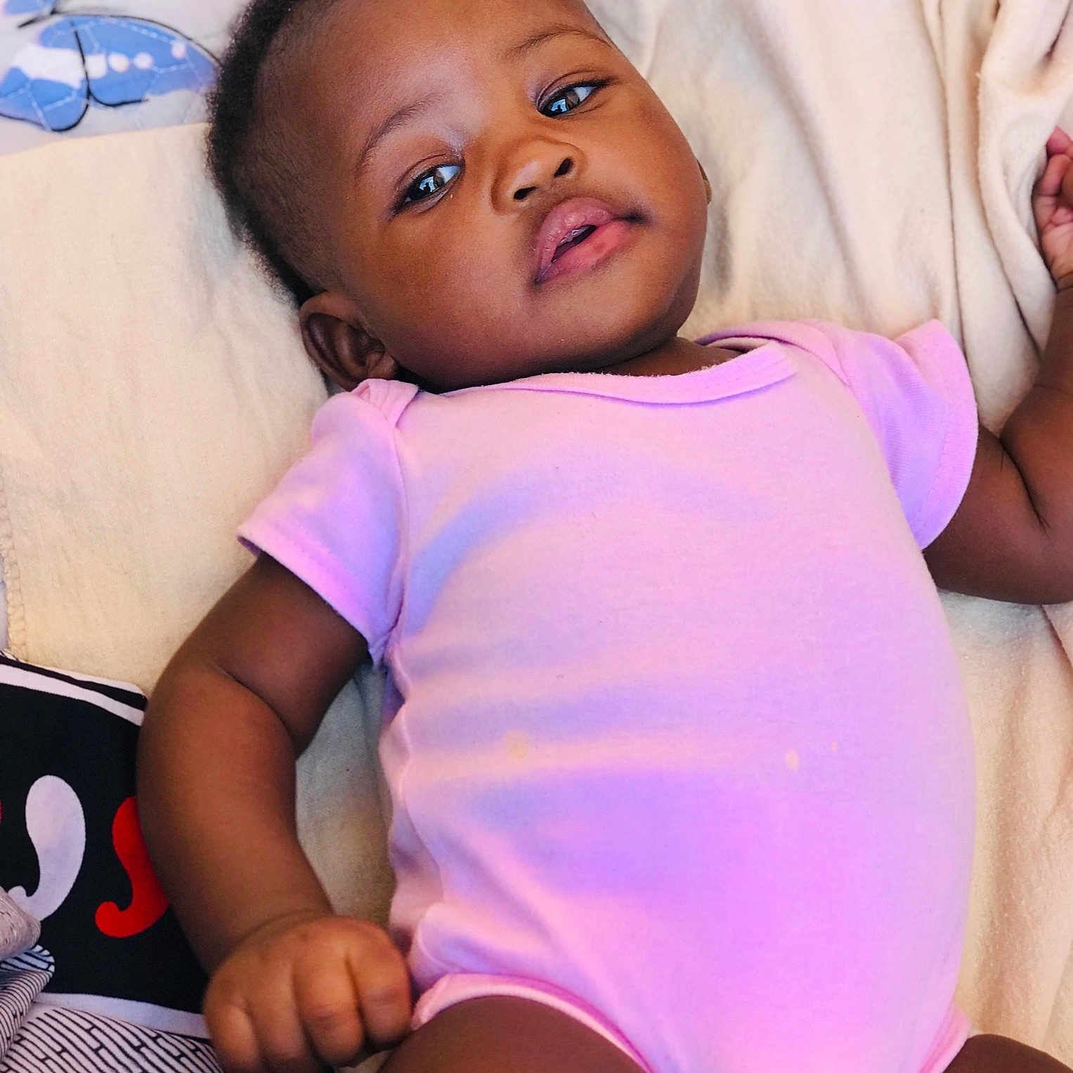 Betive is registered to the contest to win money with this photo: baby, infant, child, onesie, blanket, hand, pillow, butterfly, soft, lying_down, cute, curious, calm, indoors, person, skin, face, arm, leg, clothing