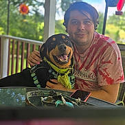 Bear joined the competition — help win amazing prizes! person, dog, smiling, outdoor, porch, glass_table, green_collar, tie_dye_shirt, happy, pet, nature, trees, daylight, phone, hand, background_blur, sitting, leash, furniture, casual