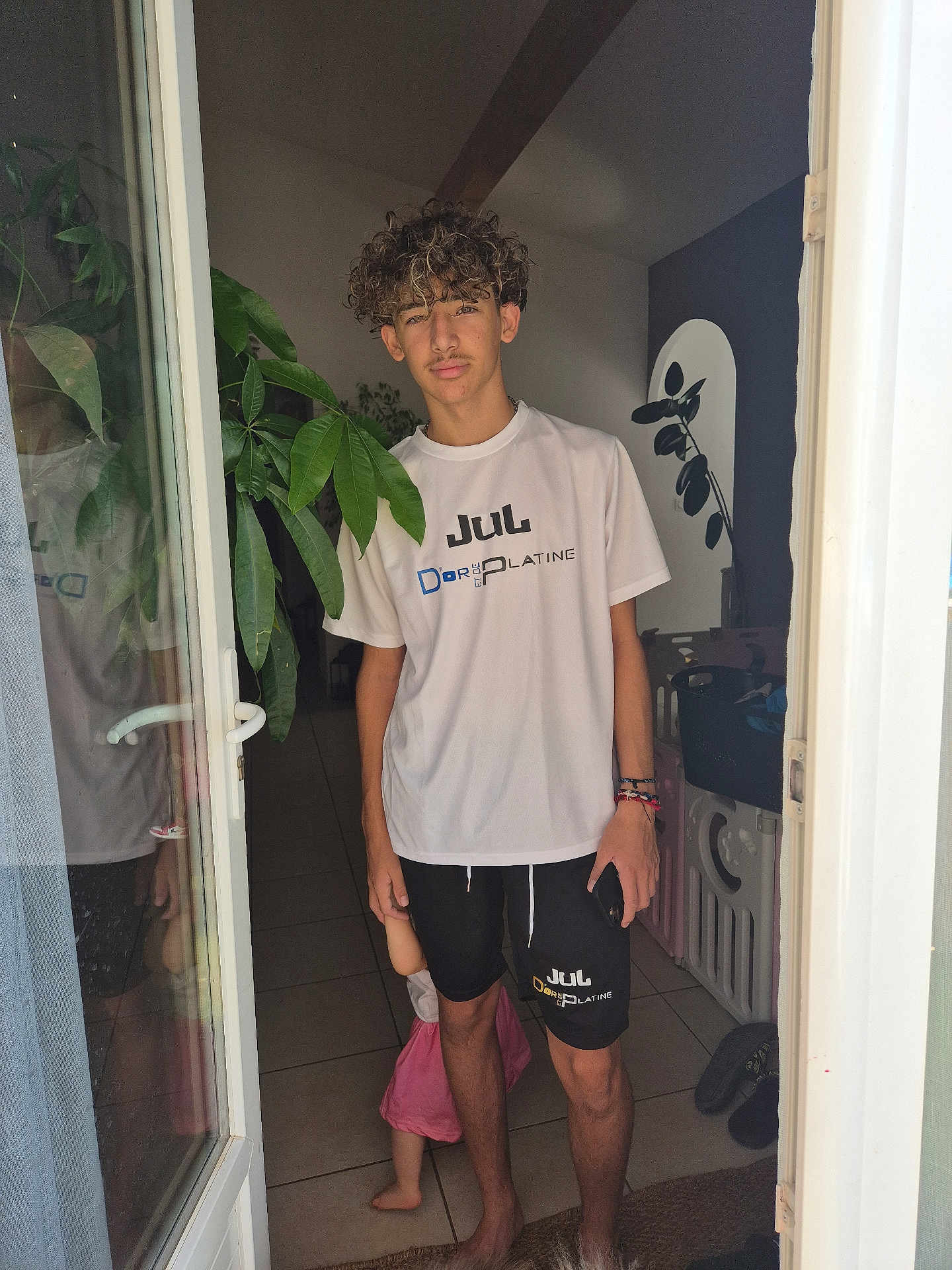 Loris participe au concours pour gagner de l'argent avec cette photo : teenager, child, doorway, curly_hair, tshirt, shorts, indoor, plant, floor_tiles, barefoot, baby_gate, reflection, bracelets, phone, shadow, window, door, pink_dress, person, casual