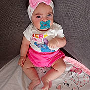 Alana a rejoint le concours — aidez-le/la à gagner de superbes lots ! baby, pacifier, headband, pink_bow, shirt, shorts, blanket, cartoon_bear, couch, gray_fabric, infant, child, cute, sitting, indoors, portrait, soft_lighting, clothing, person, face