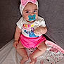 baby, pacifier, headband, pink_bow, shirt, shorts, blanket, cartoon_bear, couch, gray_fabric, infant, child, cute, sitting, indoors, portrait, soft_lighting, clothing, person, face